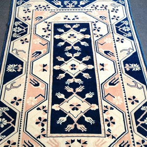 May include: A rectangular rug with a central navy blue panel featuring a diamond and floral pattern. The rug has a cream-colored background with pink and navy blue geometric borders and floral accents. The rug is a decorative home decor item.