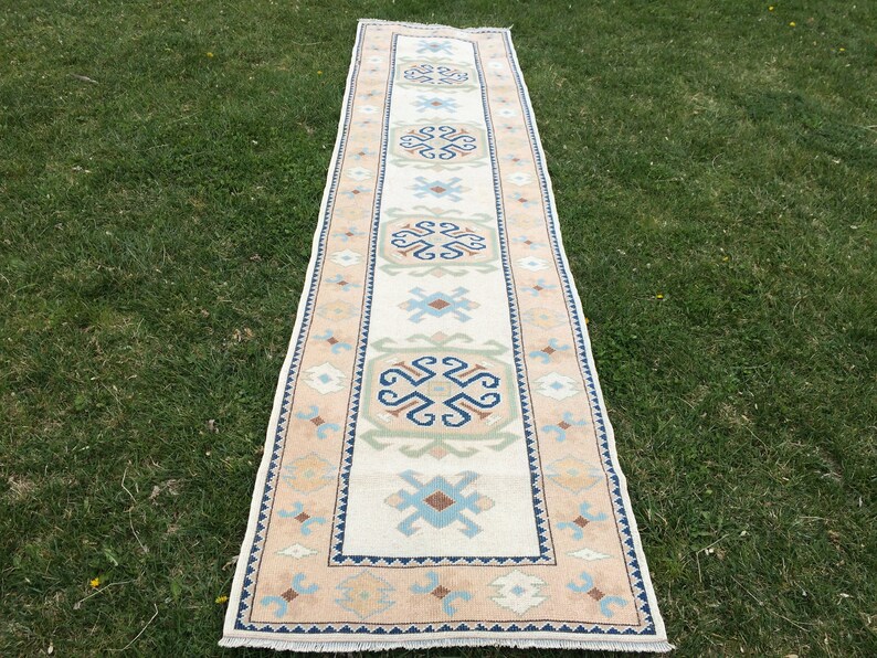 May include: A rectangular runner rug with a cream-colored central field and a border of tan, blue, and green geometric patterns. The rug is laid out on a grassy surface, showcasing its design and colors.