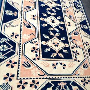 May include: A rectangular rug with a central navy blue panel featuring floral and geometric patterns. The rug has a cream-colored background with pink and navy blue borders and accents. The rug's design includes stylized floral and geometric motifs.