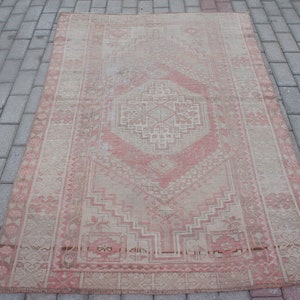 May include: A rectangular, vintage-style rug with a central geometric design in shades of pink and beige. The rug features intricate patterns and a distressed appearance, suggesting age and use. The rug is placed on a brick surface.
