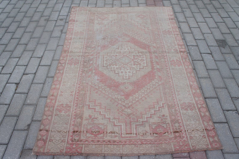 May include: A rectangular, vintage-style rug with a central geometric design in shades of pink and beige. The rug features intricate patterns and a distressed appearance, suggesting age and use. The rug is placed on a brick surface.