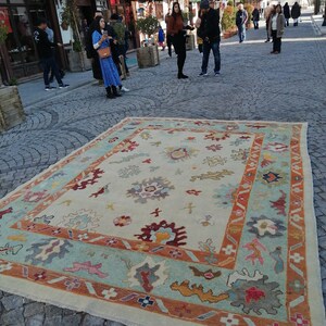 May include: A large, rectangular rug with an intricate floral design. The rug features a cream-colored central field with a border of light blue, orange, and other colors. The rug is displayed outdoors on a cobblestone street.