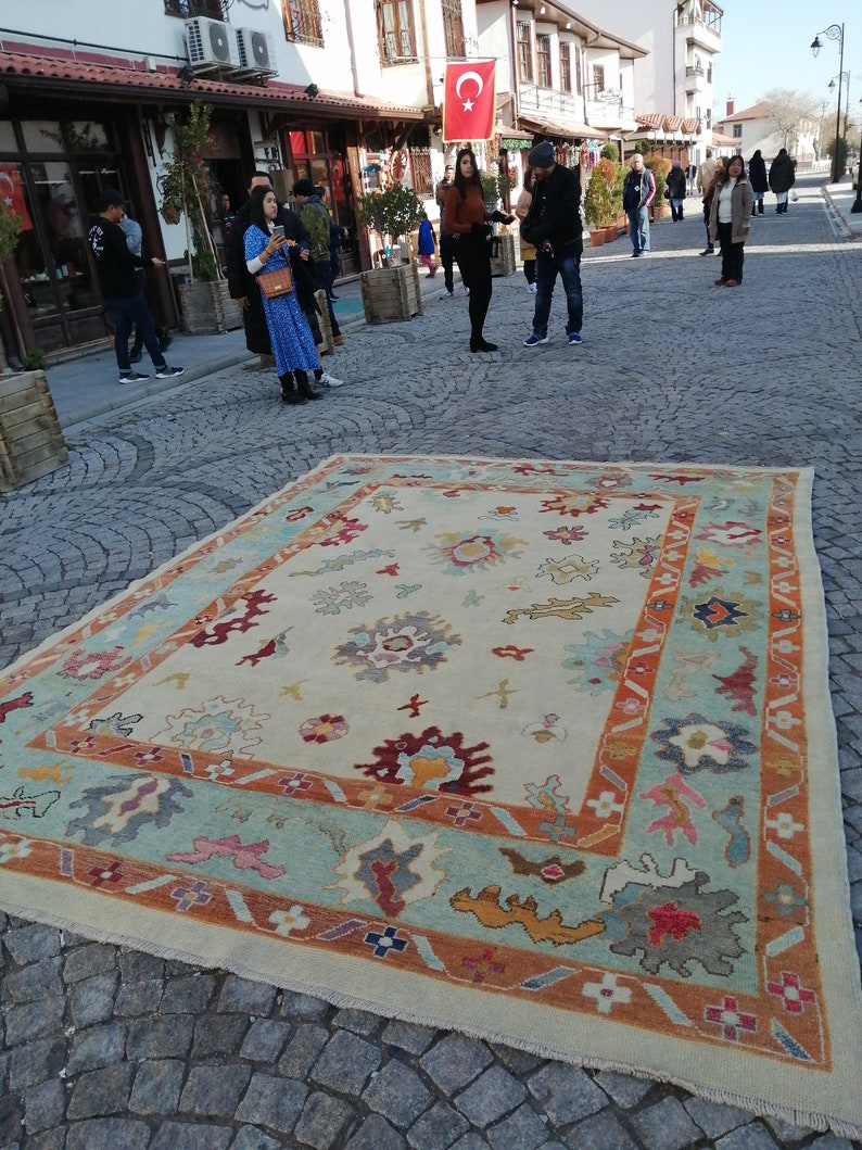 May include: A large, rectangular rug with an intricate floral design. The rug features a cream-colored central field with a border of light blue, orange, and other colors. The rug is displayed outdoors on a cobblestone street.