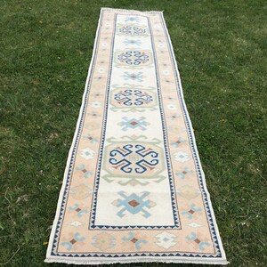 May include: A rectangular runner rug with a cream-colored central field and a repeating geometric pattern in shades of blue, beige, and green. The border is a light peach color. The rug is displayed on green grass.