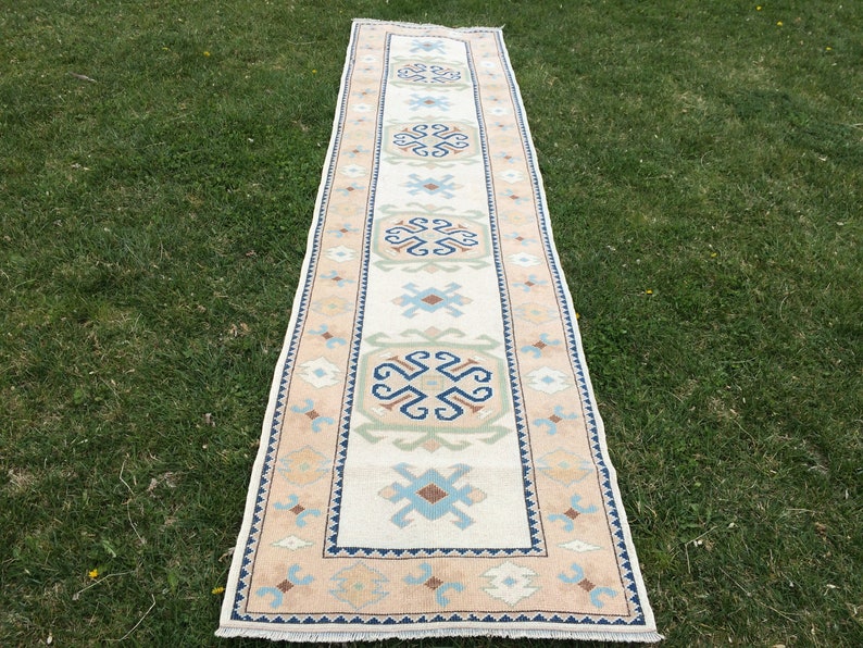 May include: A rectangular runner rug with a cream-colored central field and a repeating geometric pattern in shades of blue, beige, and green. The border is a light peach color. The rug is displayed on green grass.