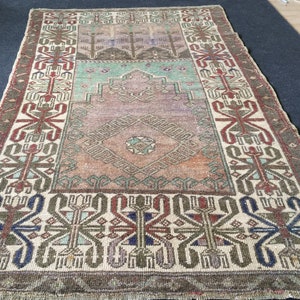May include: A rectangular vintage rug with a central diamond pattern in shades of orange, brown, and teal. The rug has a border with geometric designs in red, blue, and green on a cream background. The rug has a distressed, faded appearance.