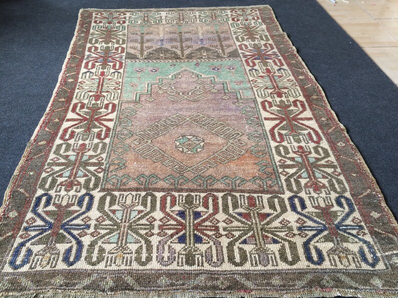 May include: A rectangular vintage rug with a central diamond pattern in shades of orange, brown, and teal. The rug has a border with geometric designs in red, blue, and green on a cream background. The rug has a distressed, faded appearance.