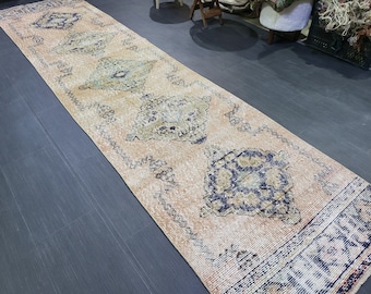 May include: A long, rectangular rug with a faded, vintage appearance. The rug features a peach-colored background with repeating geometric patterns in navy blue, olive green, and cream. The rug is likely made of woven material.