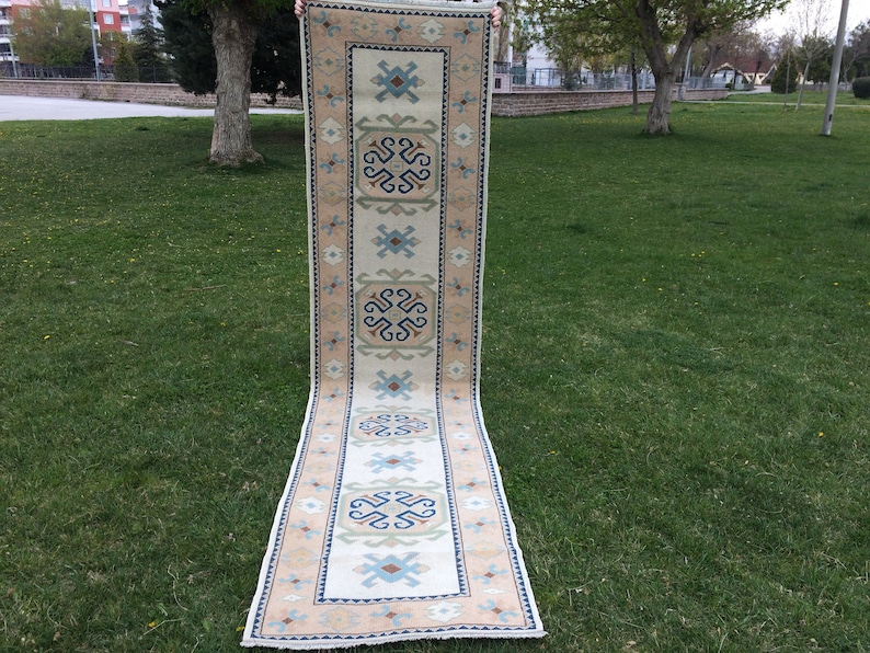 May include: A long, rectangular rug with a cream-colored central field and a patterned border. The border and central design feature geometric shapes in shades of blue, brown, and green. The rug is displayed outdoors on a grassy surface.