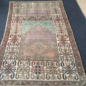 May include: Rectangular vintage rug with a central medallion and intricate geometric patterns. The rug features a colour palette of muted greens, browns, and purples, with a cream-coloured border. The rug's design includes stylised floral and geometric motifs.