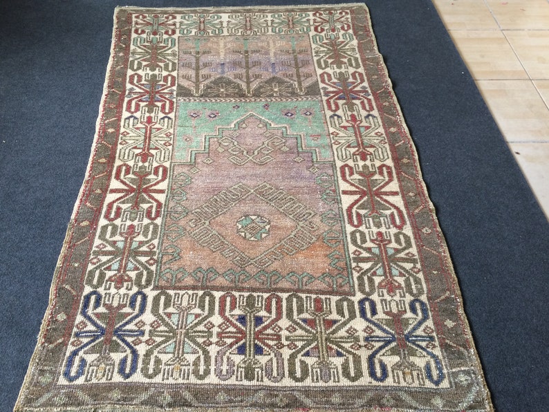 May include: Rectangular vintage rug with a central medallion and intricate geometric patterns. The rug features a colour palette of muted greens, browns, and purples, with a cream-coloured border. The rug's design includes stylised floral and geometric motifs.