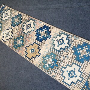 May include: A rectangular runner rug with a geometric pattern in shades of blue, white, beige, and pink. The design features repeating square and rectangular shapes with intricate details. The rug is likely made of woven fabric.