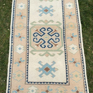 May include: A rectangular beige and cream-colored rug with a central cream field and a patterned border. The rug features geometric designs in blue, brown, and green. The rug has a fringe on the short ends.