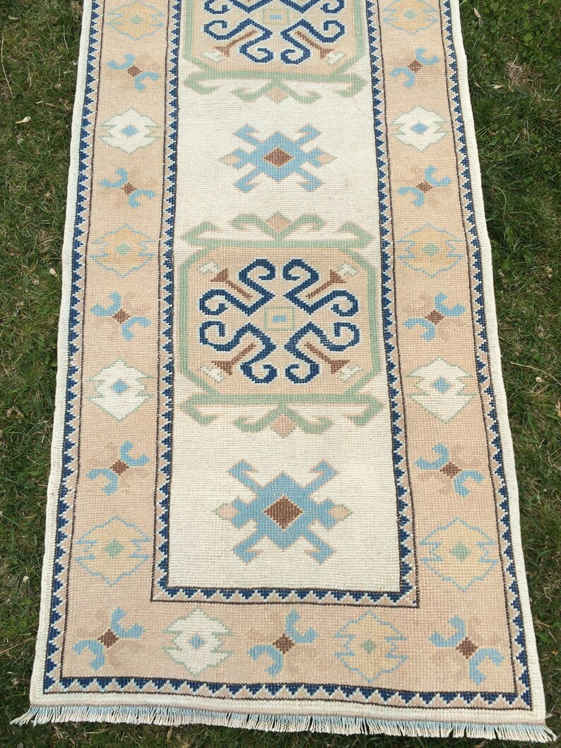May include: A rectangular beige and cream-colored rug with a central cream field and a patterned border. The rug features geometric designs in blue, brown, and green. The rug has a fringe on the short ends.