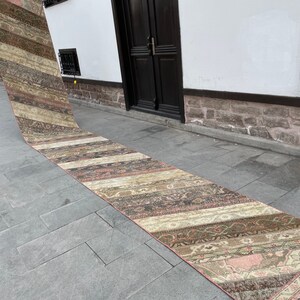 Può includere: Un lungo tappeto rettangolare con un design patchwork. Il tappeto presenta una varietà di colori, tra cui beige, marrone e rosa, con motivi intricati. Il tappeto è esposto all'aperto su una superficie in pietra.