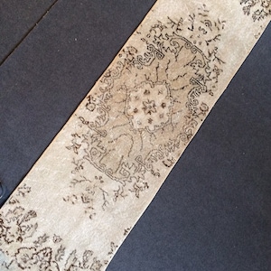 Puede incluir: Una alfombra de pasillo beige con un intrincado diseño floral marrón oscuro. El diseño de la alfombra presenta un medallón central y bordes con motivos florales repetidos. La alfombra se muestra sobre una superficie gris oscuro.