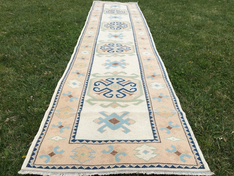 May include: A rectangular, cream-colored runner rug with a central design of geometric shapes in blue, brown, and green. The border is a light peach color with a repeating pattern, and the rug is placed on green grass.