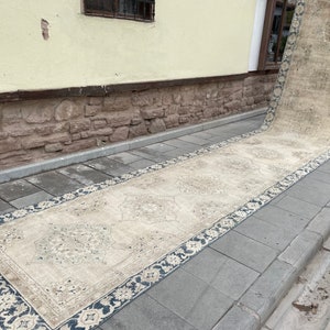 May include: A long, rectangular beige rug with a dark blue border featuring floral patterns. The rug is placed on a gray stone surface, with a building and window in the background. The rug has a distressed, vintage appearance.