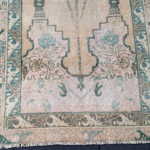 May include: A vintage rug with a central design in shades of peach and beige, framed by a border of teal and brown. The rug features floral motifs and geometric patterns, showcasing a distressed, antique aesthetic.