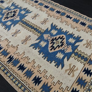 Puede incluir: Alfombra crema con un patrón geométrico en azul, beige y melocotón. La alfombra presenta un diseño central con formas repetidas y un borde con un patrón similar. La alfombra es rectangular y parece estar hecha de material tejido.