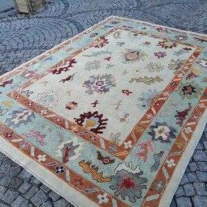 May include: Rectangular area rug with a cream-colored field and a border in shades of teal, rust, and brown. The rug features floral and geometric patterns in various colors, including burgundy, yellow, and blue. The rug is placed on a cobblestone surface.