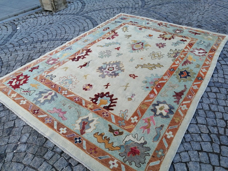 May include: Rectangular area rug with a cream-colored field and a border in shades of teal, rust, and brown. The rug features floral and geometric patterns in various colors, including burgundy, yellow, and blue. The rug is placed on a cobblestone surface.