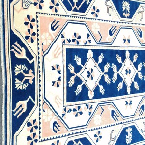 May include: Close-up of a rectangular rug with a detailed geometric pattern. The rug features a navy blue border and central design, with cream and light pink accents. Floral and geometric shapes are repeated throughout the design, creating a traditional aesthetic.