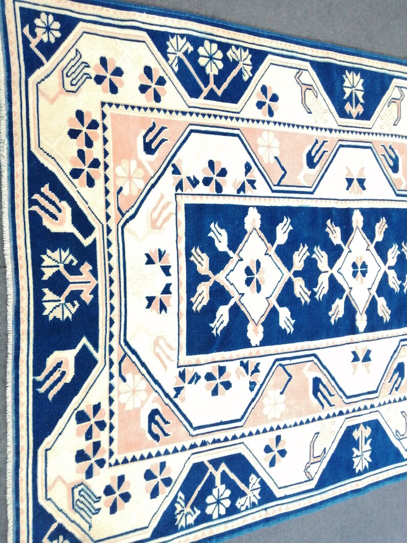 May include: Close-up of a rectangular rug with a detailed geometric pattern. The rug features a navy blue border and central design, with cream and light pink accents. Floral and geometric shapes are repeated throughout the design, creating a traditional aesthetic.