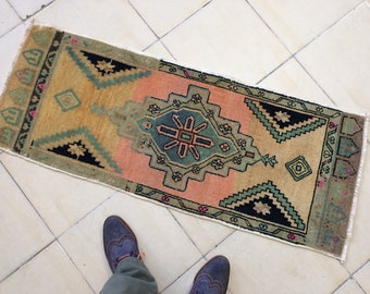 May include: A rectangular, patterned rug with a central design in shades of pink, green, and blue. The rug has a beige background with geometric shapes and a fringed edge. The rug is on a tiled floor.