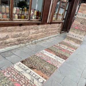 May include: A long, rectangular runner rug with a patchwork design. The rug features various patterns and colors, including pink, brown, beige, and green. The rug is displayed outdoors on a stone walkway.