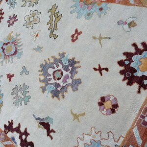 May include: Cream-colored area rug with a detailed floral pattern in shades of burgundy, blue, and gold. The rug features a border with geometric designs and a soft, textured appearance. The design includes stylized floral and abstract shapes.
