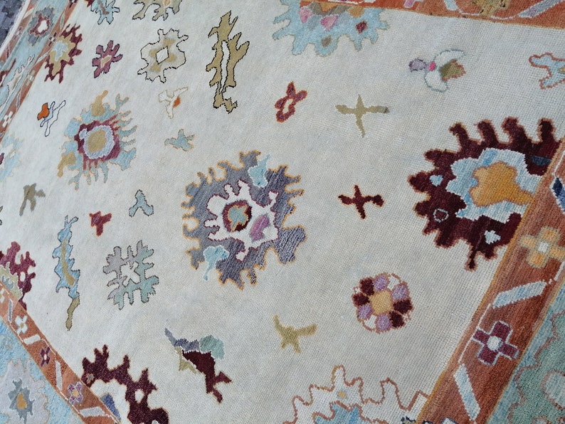 May include: Cream-colored area rug with a detailed floral pattern in shades of burgundy, blue, and gold. The rug features a border with geometric designs and a soft, textured appearance. The design includes stylized floral and abstract shapes.