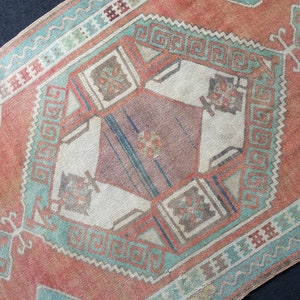 Puede incluir: Una alfombra antigua con un dise&ntilde;o geom&eacute;trico. La alfombra presenta una forma de diamante central en tonos de rosa, marr&oacute;n y crema, rodeada de bordes turquesa y naranja. El patr&oacute;n de la alfombra incluye motivos geom&eacute;tricos repetidos.