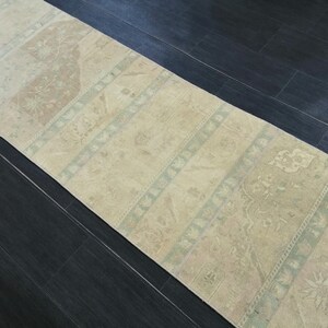 Peut inclure: Un tapis rectangulaire de style vintage, aux tons beige et marron. Le tapis présente un motif répétitif de rayures et de motifs floraux, avec des accents bleu clair et vert. Il est posé sur un sol en bois foncé.