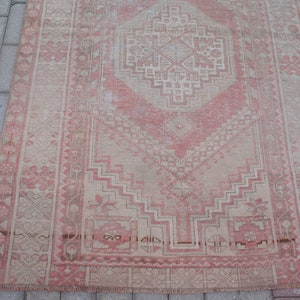 May include: A rectangular, distressed rug with a central diamond pattern in shades of pink and beige. The rug features geometric designs and floral motifs, with a faded, vintage appearance. The rug is placed on a gray brick surface.