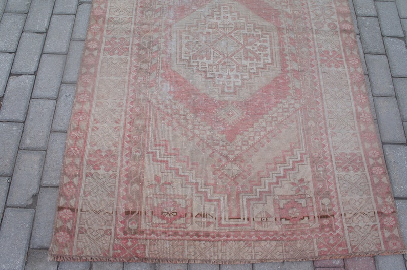 May include: A rectangular, distressed rug with a central diamond pattern in shades of pink and beige. The rug features geometric designs and floral motifs, with a faded, vintage appearance. The rug is placed on a gray brick surface.