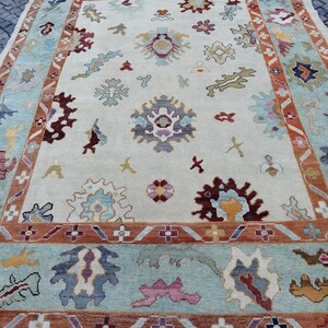 May include: A large, rectangular area rug with an intricate floral pattern. The rug features a cream-colored central field with scattered floral motifs in shades of blue, burgundy, and gold. A border of light blue and rust-colored bands frames the design.