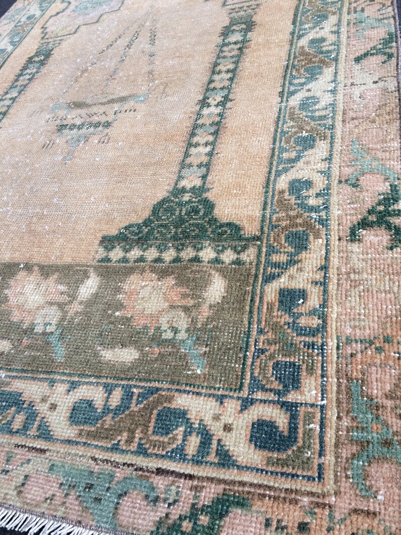 May include: A vintage rug with a central peach field and a dark green border. The border features a repeating geometric pattern in teal and brown. The rug has a distressed appearance, with faded colors and visible wear.