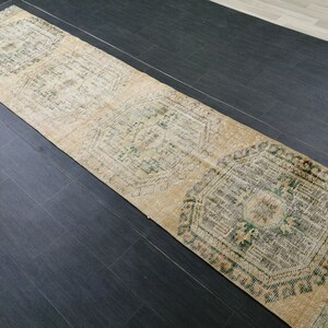 May include: A long, rectangular runner rug with a distressed, vintage appearance. The rug features a beige background with repeating geometric patterns in shades of green and brown. The rug is placed on a dark wood floor.
