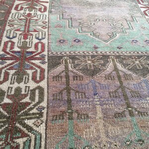 May include: A distressed vintage rug with a patchwork design. The rug displays geometric patterns in brown, green, purple, and beige. The border features a detailed, symmetrical design with red and blue accents. The rug has a worn, aged appearance.