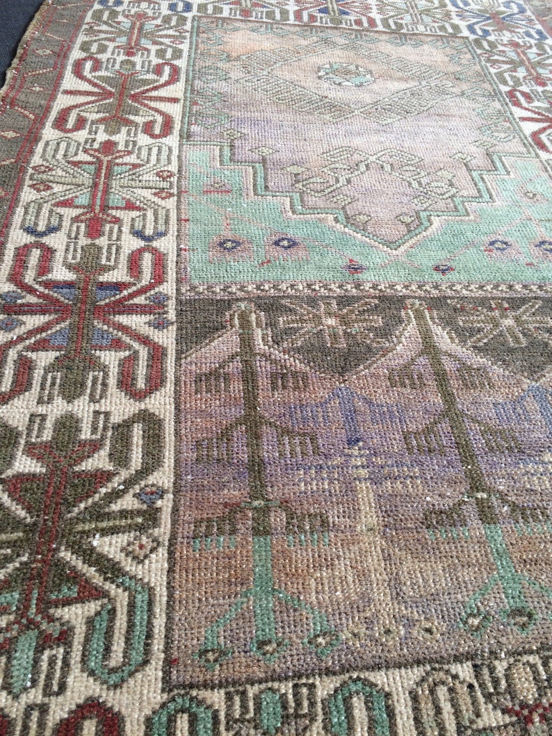 May include: A distressed vintage rug with a patchwork design. The rug displays geometric patterns in brown, green, purple, and beige. The border features a detailed, symmetrical design with red and blue accents. The rug has a worn, aged appearance.