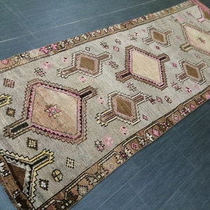May include: A rectangular, vintage-style rug with a neutral gray background and geometric patterns. The rug features diamond and rectangular shapes in shades of brown, pink, and beige, with a detailed border. The rug is suitable for a hallway or living room.
