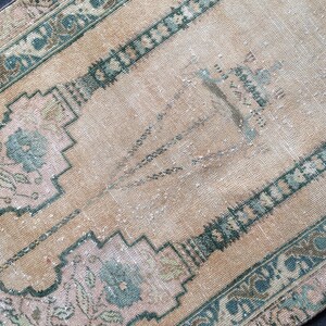 May include: A rectangular, vintage-style rug with a faded, distressed appearance. The rug features a central field in a warm beige hue, framed by a border of teal and pink floral patterns. The design includes geometric shapes and floral motifs.