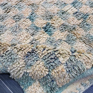 Op de afbeelding: Close-up van een getextureerd tapijt met een geometrisch patroon. Het tapijt heeft een combinatie van cr&egrave;me, lichtblauw en beige kleuren. Het tapijt heeft een dikke, zachte pool en een geweven rand.