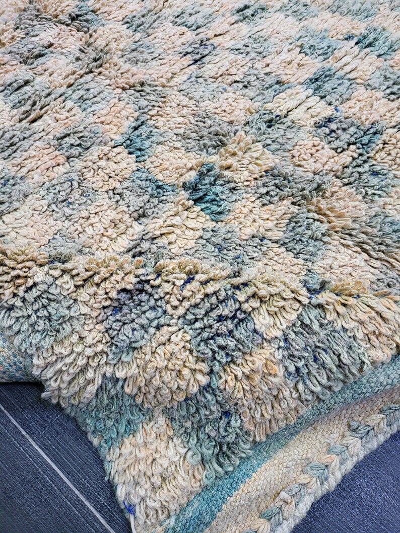 Op de afbeelding: Close-up van een getextureerd tapijt met een geometrisch patroon. Het tapijt heeft een combinatie van cr&egrave;me, lichtblauw en beige kleuren. Het tapijt heeft een dikke, zachte pool en een geweven rand.