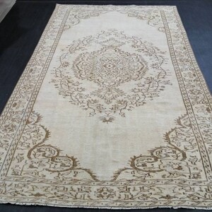 Puede incluir: Una alfombra rectangular de color crema con un intrincado patrón floral marrón. La alfombra presenta un diseño de medallón central y un borde detallado. La alfombra está colocada sobre una superficie oscura, mostrando su diseño.
