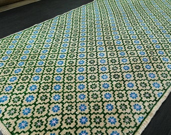 May include: A rectangular rug with a repeating pattern of green and blue floral designs on a cream background. The rug has a fringed edge and is likely made of woven fabric.