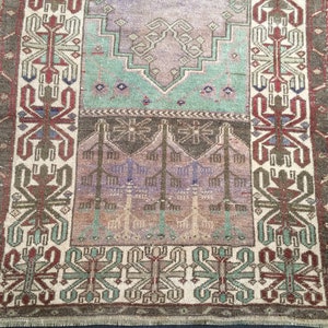 May include: A rectangular, vintage rug with a faded, distressed appearance. The rug features a central design in shades of purple, green, and brown, with a border in similar tones. The pattern includes geometric and floral motifs.