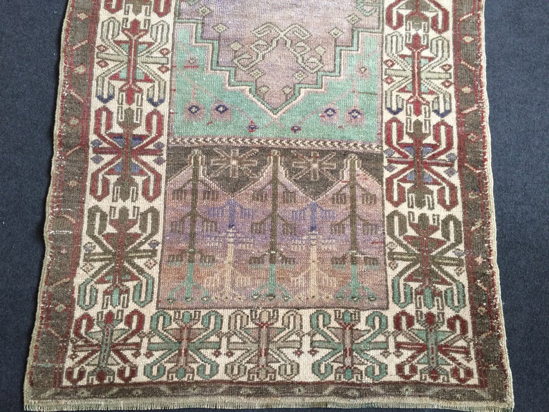 May include: A rectangular, vintage rug with a faded, distressed appearance. The rug features a central design in shades of purple, green, and brown, with a border in similar tones. The pattern includes geometric and floral motifs.
