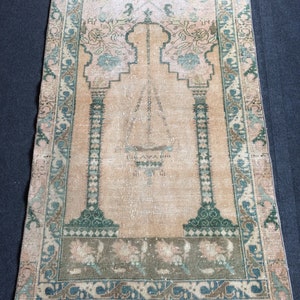 May include: A rectangular, vintage prayer rug with a central arch design. The rug features a peach-colored field with green and blue floral accents. The border is a mix of green, blue, and brown patterns, with a fringed edge.